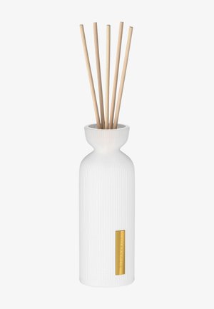 Vit ribbad keramikdiffusor med en avsmalnande hals, prydd med fem bambupinnar och en guldfärgad etikett. Slät textur och minimalistisk design.