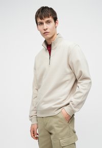 Junger Mann in einem beigen Sweatshirt mit Viertelreißverschluss über einem roten Shirt und hellbraunen Cargo-Hosen, der mit einer Hand in der Tasche steht.