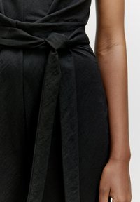 Schwarzes Kleid aus Stoff mit gebundenem Taillendetail, glatter Textur und tailliertem Oberteil, mit Fokus auf dem Knoten und den Drapierungen.