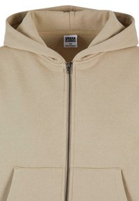 Béžová mikina na zips vyrobená z mäkkej tkaniny, s klokankou a nastaviteľnou kapucňou. Obsahuje strieborný zips a prešívané detaily.
