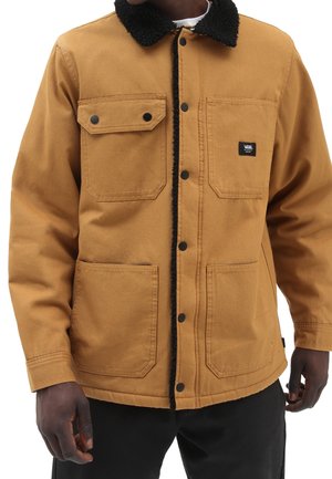 Mężczyzna ubrany w jasnobrązową kurtkę na guziki z czarnym kołnierzem typu sherpa i czterema przednimi kieszeniami, z których jedna ma naszywkę z logo Vans.