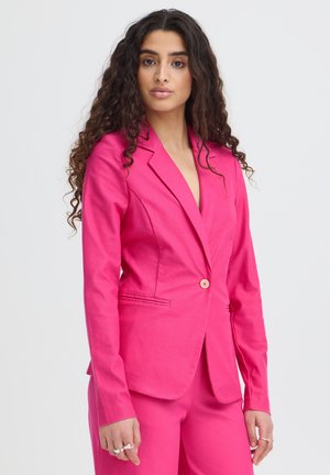 Vrouw met lang krullend haar die een aansluitend felroze colbert en bijpassende broek draagt, staande tegen een effen achtergrond.