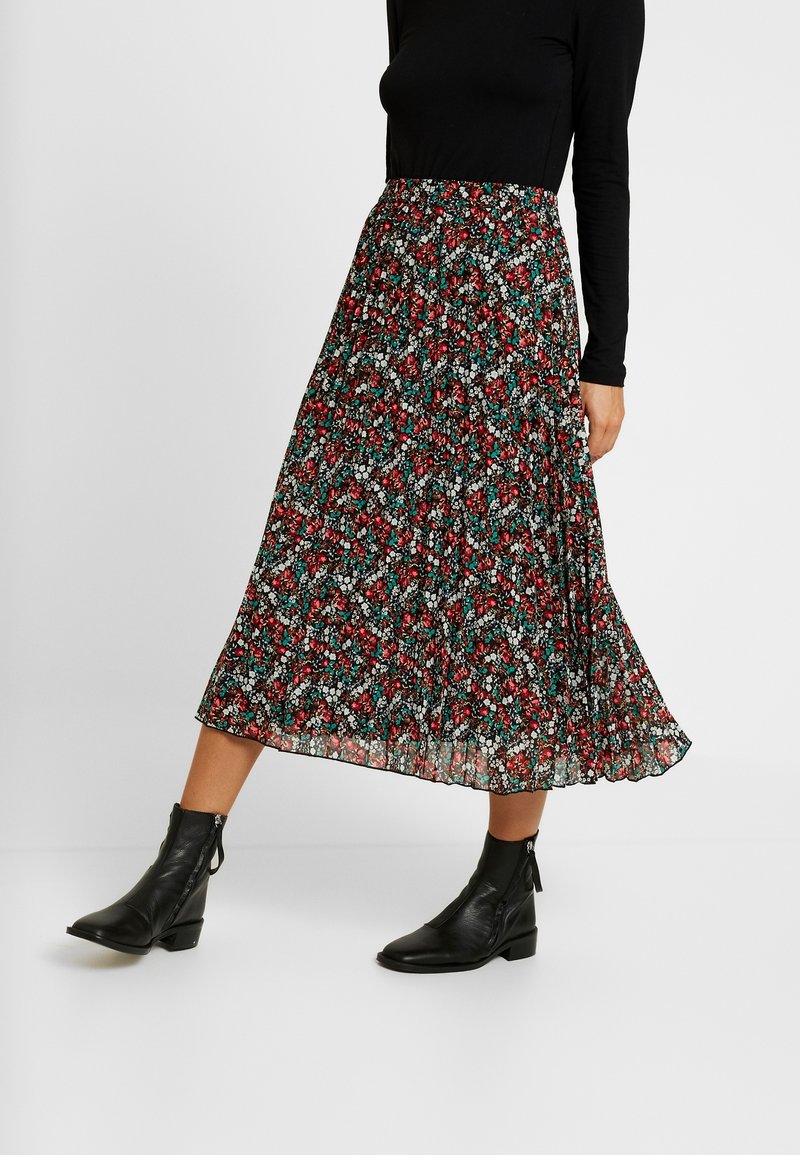 Femme portant une jupe midi plissée avec un motif floral rouge, blanc et sarcelle, un haut à manches longues noir et des bottines noires.