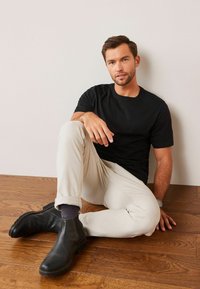 Camiseta negra de manga corta, pantalones ajustados beige, botas de ante negras hasta el tobillo y calcetines oscuros con estampado, colocados sobre un suelo de madera.