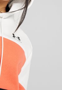 Sudadera blanca y naranja con cordón. Presenta el logo de Under Armour en el pecho y una tela texturizada con un diseño en bloques de color.