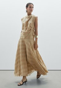 Beiges kariertes Kleid mit lockerem Schnitt, ärmellosem Design und seitlichen Taschen; kombiniert mit schwarzen Riemchen-Heels für einen ausgewogenen Look.