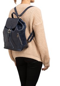 Zaino in pelle blu navy con chiusura a coulisse, due cerniere laterali e spallacci regolabili, indossato sopra un maglione di maglia color crema.