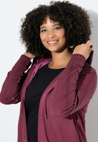 Burgunderfarvet hoodie med lynlås og tekstureret finish, med hætte og sidelommer, båret over en sort top, præsenteret mod en hvid baggrund.