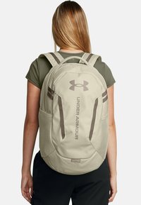 Under Armour kuprinė, smėlio spalvos audinys, ergonomiškas dizainas, paminkštinti diržai, šoniniai tinkliniai kišenės, iškilmuotas logotipas, tvirtas siuvimas ir lygus paviršius.