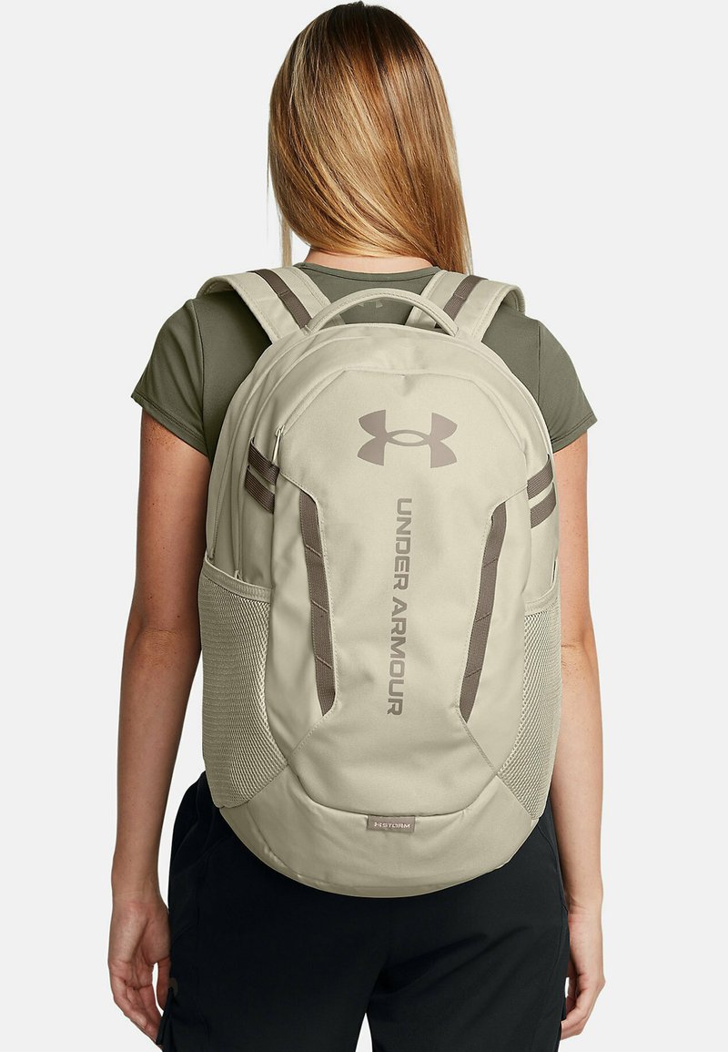 Under Armour kuprinė, smėlio spalvos audinys, ergonomiškas dizainas, paminkštinti diržai, šoniniai tinkliniai kišenės, iškilmuotas logotipas, tvirtas siuvimas ir lygus paviršius.