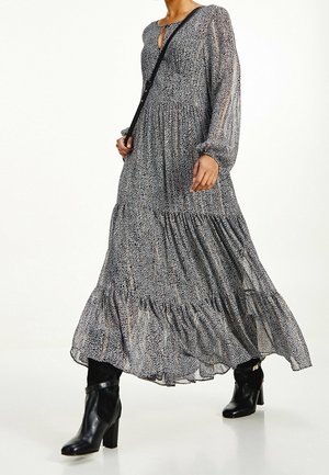 Femme portant une longue robe noire et blanche à motifs avec manches longues, des bottes noires à talons et un sac bandoulière, marchant vers l'avant.
