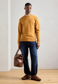 Felpa girocollo di cotone color senape con polsini a costine, abbinata a jeans blu scuro e borsa a tracolla in pelle marrone. Sneakers in suede marroni.