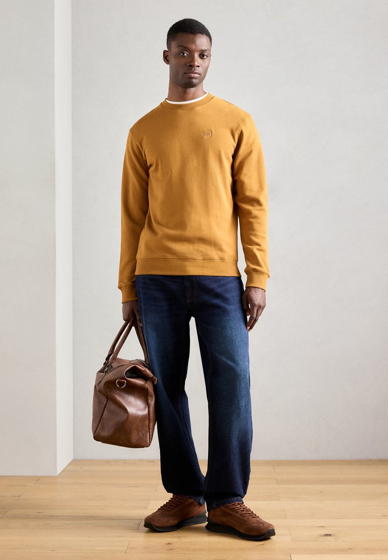 Felpa girocollo di cotone color senape con polsini a costine, abbinata a jeans blu scuro e borsa a tracolla in pelle marrone. Sneakers in suede marroni.