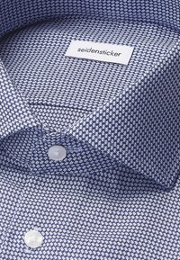 Close-up van een gevouwen blauw-wit gestreept overhemd met witte knopen en een "seidensticker" label aan de binnenkant van de kraag.