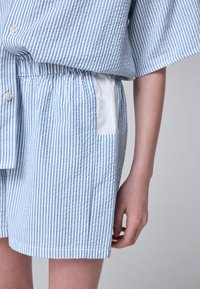 Plava i bijela košulja s okomitim prugama, kratkih rukava, s prednjim gumbima, elastičnim pojasom i bijelim džepom od tkanine.
