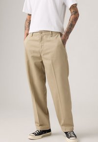 Beige, wide-leg trousers with a flat front, two side pockets, and belt loops, paired with black sneakers and a plain white t-shirt.