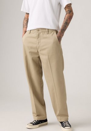 Pantalons beige à jambes larges avec un devant plat, deux poches latérales et des passants pour ceinture, associés à des baskets noires et un t-shirt blanc uni.
