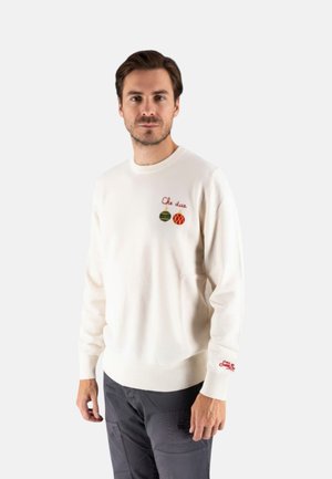 Uomo che indossa un maglione bianco con testo rosso e decorazioni natalizie, abbinato a pantaloni scuri, in piedi di fronte a uno sfondo semplice.