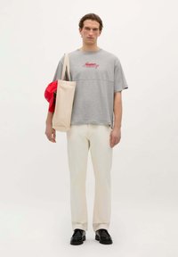 Grijze T-shirt met korte mouwen en rode tekst, crèmekleurige rechte broek en zwarte schoenen; draagt een beige tote bag met een rode pet erin.