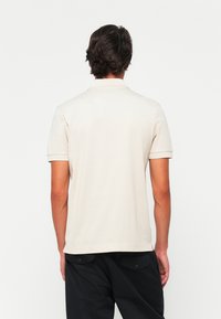 Pikétrøye i lys beige med korte ermer, krage og rett fald. Har en glatt tekstur og et minimalt design, uten synlige mønstre.