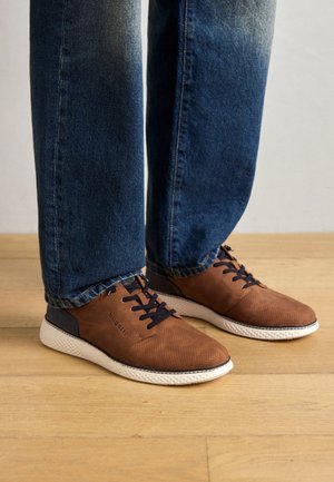 Sneaker in pelle marrone forate con accenti blu navy, suola bianca e lacci neri, abbinate a jeans blu sbiaditi. Sfondo in parquet.