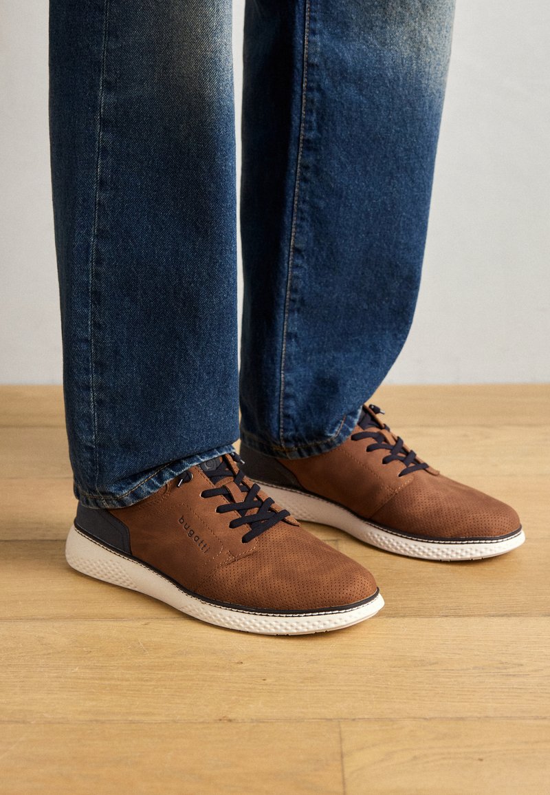Baskets perforées marron avec des accents bleu marine, semelle blanche et lacets noirs, associées à un jean bleu délavé. Fond en parquet en bois.