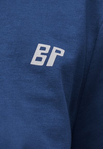 Μπλε ύφασμα με άσπρα στιλιζαρισμένα γράμματα "BP" τυπωμένα, που δείχνουν υφή και ελαφρές πτυχώσεις στο υλικό.