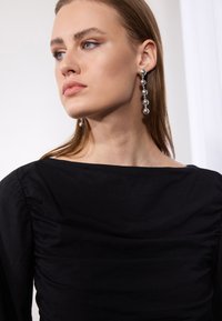 Zwarte stoffen top met een brede halslijn en geplooide accenten. Model draagt lange zilveren oorbellen met ronde kralen, voorzien van een gladde afwerking.