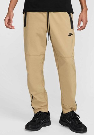 Pantalons de jogging beiges avec des accents latéraux noirs, taille avec cordon de serrage et poches zippées. Portés avec des chaussures de sport noires présentant un design texturé.