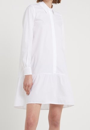 Kobieta w długiej koszuli sukience z długim rękawem, kołnierzykiem i falbanami na dole, stojąca na jednolitym jasnym tle.