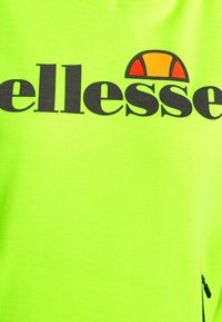 Camiseta verde brillante con gran logotipo negro de "ellesse" y un símbolo de acento multicolor. Textura suave con un detalle de cremallera lateral.