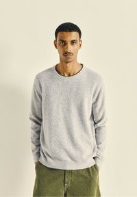Pull léger gris clair à col rond en matériau doux, avec des côtes aux poignets et à l'ourlet, associé à un pantalon vert olive. Design minimaliste.
