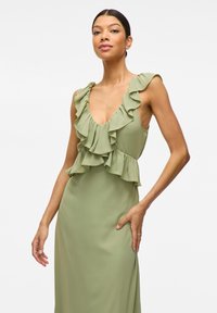 Rochie verde fără mâneci, cu un decolteu adânc în V și detalii cu volane stratificate la gât și talie. Material neted și fluid.