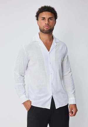 Homme aux cheveux courts et bouclés avec une barbe, portant une chemise blanche à manches longues boutonnée et un pantalon noir, debout avec une main dans la poche.