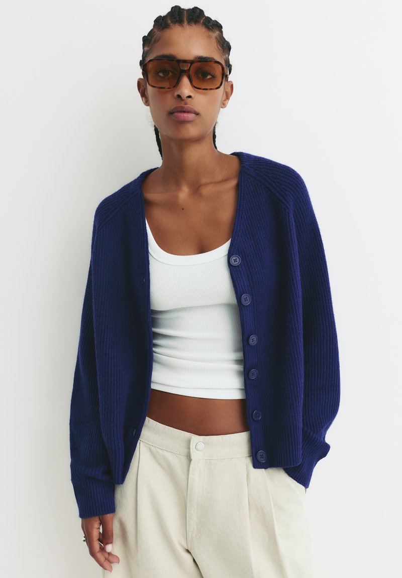 Mornarski rebrasti kardigan s gumbima sprijeda, nošen preko bijelog majice. Model ga kombinira s bež hlačama; sadrži suptilnu teksturu i opušteni kroj.