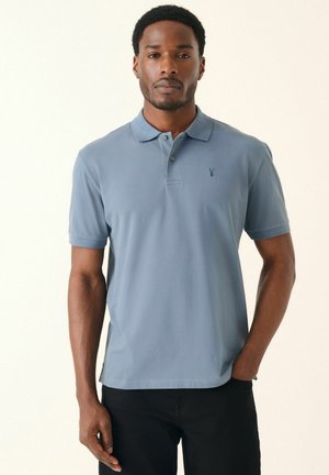 Lichtblauw polo shirt met een slim fit, korte mouwen, een knoopsluiting met twee knopen en een klein geborduurd logo op de borst. Zwarte broek wordt gedragen.