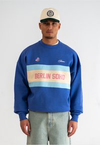 Sweatshirt albastru cu inscriptionarea "BERLIN SOHO" în dungi roz și crem, având manșete ribetite și guler rotund. Căciulă cu partea din față crem și brăc de culoare verde.