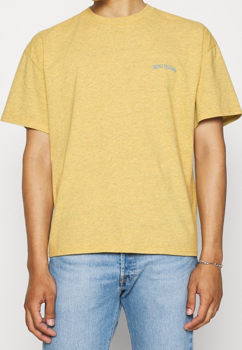 Gul kortärmad t-shirt i mjuk bomull, med en diskret blå logotyp på vänster bröstområde. Klassisk rund halsringning, avslappnad passform.