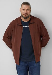 Brązowa kurtka na zamek z teksturowanym wzorem; prążkowany kołnierz i mankiety; nałożona na granatową koszulę. Noszona z niebieskimi jeansami.
