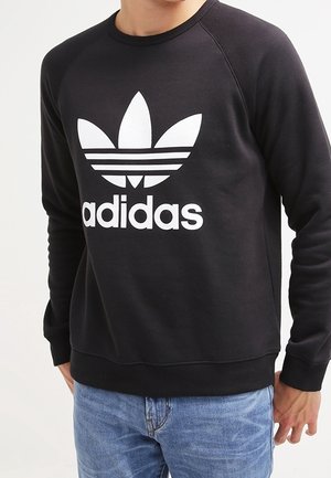 Osoba ubrana w czarną bluzę Adidas z białym logo i napisem, w połączeniu z niebieskimi dżinsami, stoi na tle jednolitego tła.