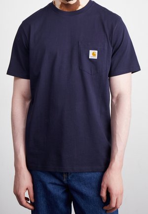 Homme portant un t-shirt bleu marine à manches courtes avec poche poitrine et patch logo Carhartt, associé à un jean bleu, sur fond uni.
