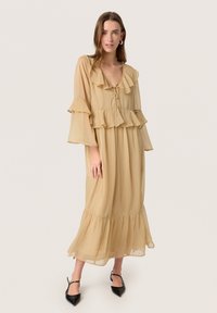 Beige dlouhé šaty s dlouhými rukávy a volánky kolem výstřihu a rukávů, splývavý silueta a zúžený lem. Nosené s černými špičatými podpatky.