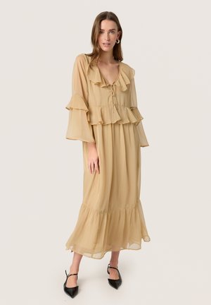 Robe longue beige à manches longues avec des volants au niveau du col et des manches, silhouette fluide et ourlet fuselé. Portée avec des talons noirs à bouts pointus.