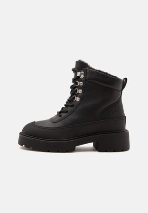 Bottine noire en cuir à lacets avec une semelle épaisse, des œillets métalliques, un soufflet de tirage et une doublure en fourrure visible au niveau du col.