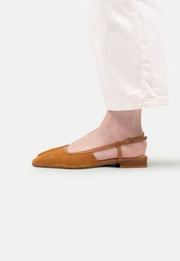 Vedere laterală a unui picior purtând o sandală din suede bej cu toc mic, asociată cu pantaloni albi scurți.