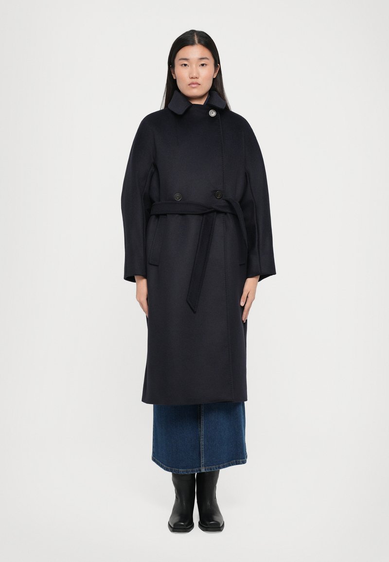 Long navy coat made of a soft, textured material, featuring a high collar, double buttons, and a matching belt. Worn over a denim skirt.