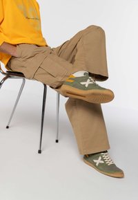 Sneaker verde con accenti gialli e bianchi, tessuto testurizzato e suola in gomma. Indossati con pantaloni cargo beige e una camicia oversize gialla.