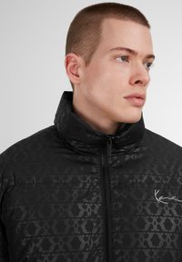 Giacca nera imbottita con colletto alto, caratterizzata da un motivo geometrico tono su tono, zip frontale e dettagli firmati discreti sul petto.