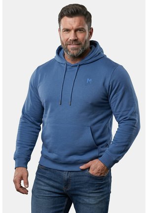 Mann trägt einen blauen Hoodie mit Fronttasche und blaue Jeans, steht mit einer Hand in der Tasche vor einfachem Hintergrund.