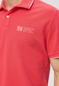 Czerwona koszulka polo z teksturowanej tkaniny, z jasnoróżowym kołnierzykiem i haftowanym logo na piersi. Klasyczny krój z zapięciem na guziki.